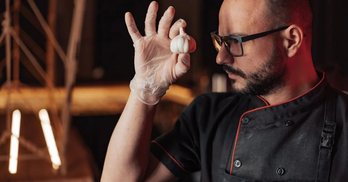 Chef chauve portant des lunettes, tenant de l'ail avec des gants dans une cuisine.