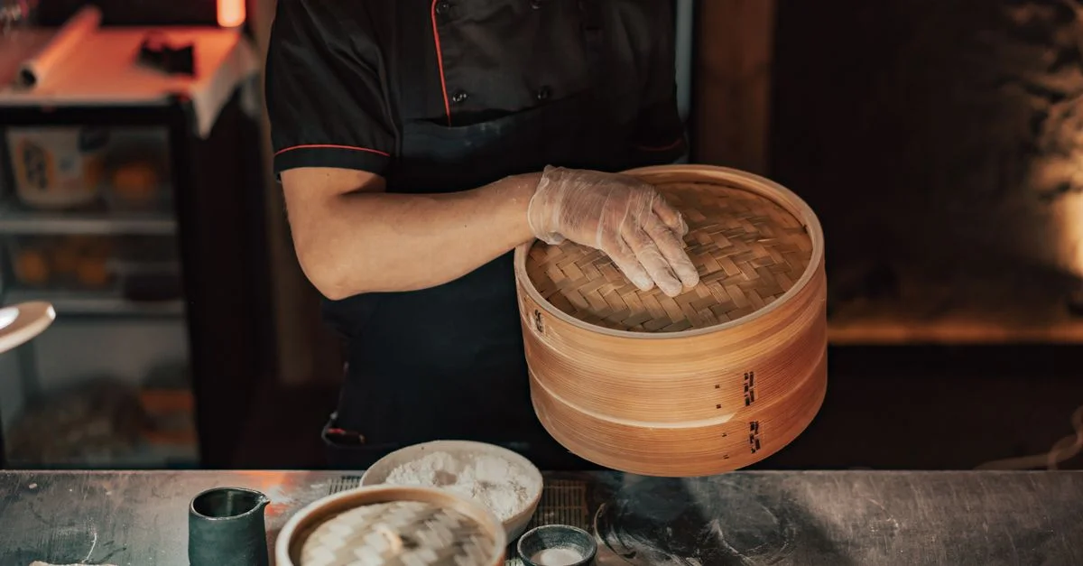 Chef en uniforme utilisant un panier vapeur en bambou pour la préparation de plats asiatiques dans une cuisine de restaurant.