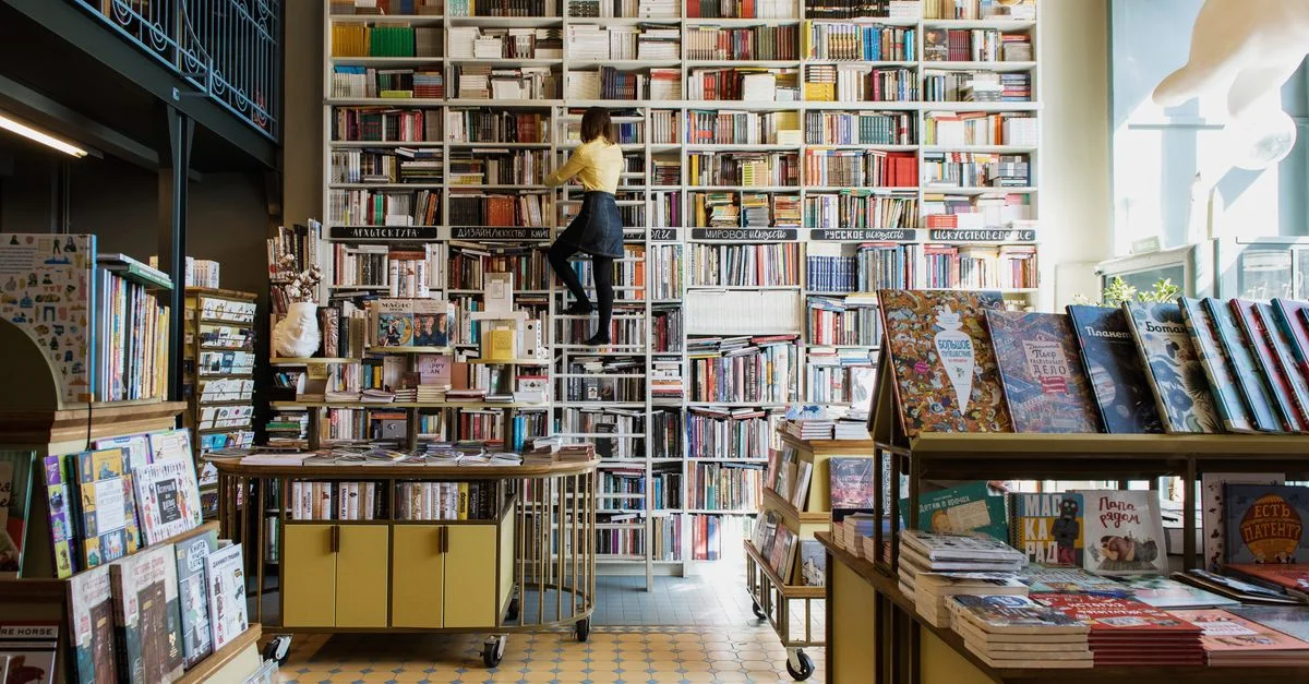 Une personne monte une échelle pour atteindre un livre dans une charmante librairie bien éclairée remplie de titres divers.