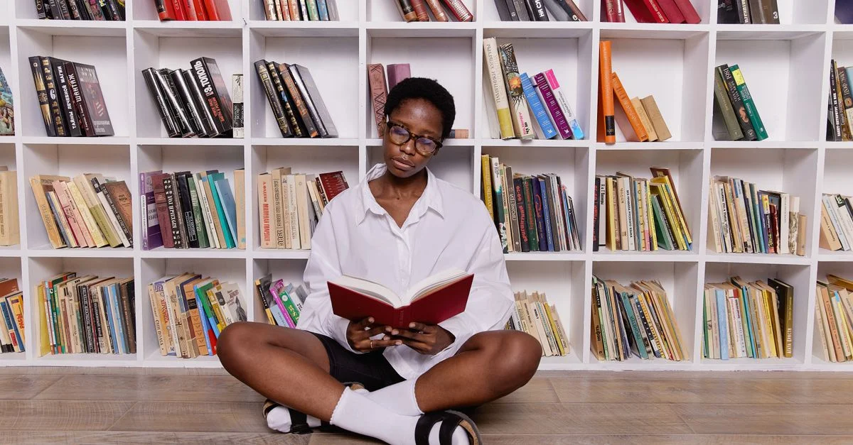 Femme noire assise par terre en lisant un livre dans une bibliothèque moderne avec des étagères blanches remplies de livres.