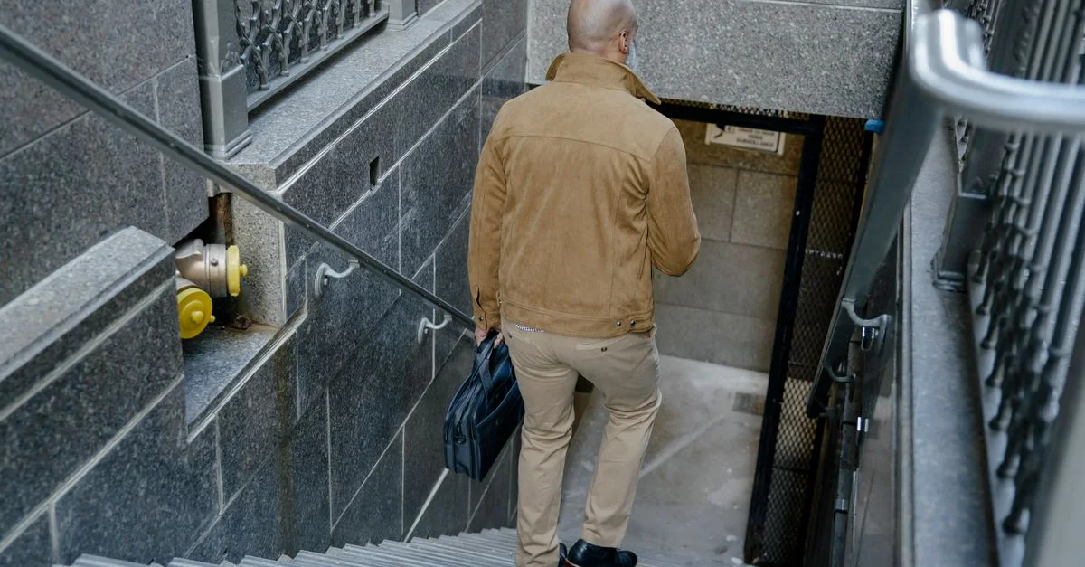 Un homme chauve en manteau beige descend des escaliers en portant une mallette dans un cadre urbain.