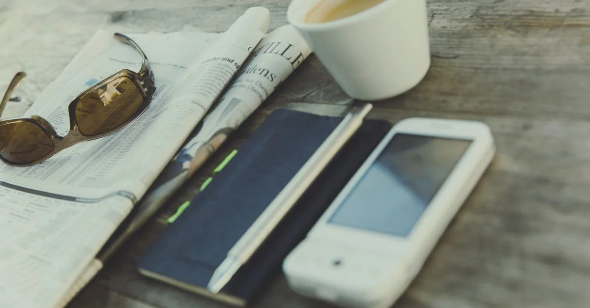 Scène relaxante avec café, journal, lunettes de soleil, carnet et smartphone sur une table en bois rustique.