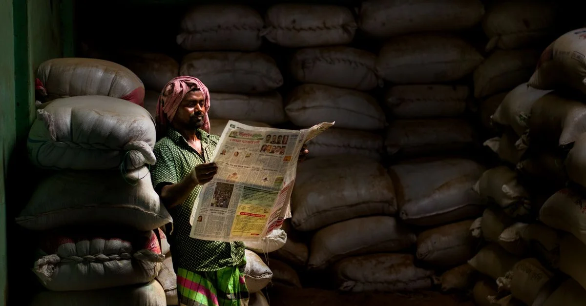Un homme en tenue traditionnelle lit un journal dans un storeroom faiblement éclairé avec des sacs empilés.
