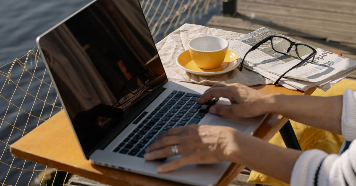 Une personne tapant sur un ordinateur portable en plein air au bord d'un lac avec un café et un journal, idéal pour des images de travail à distance.