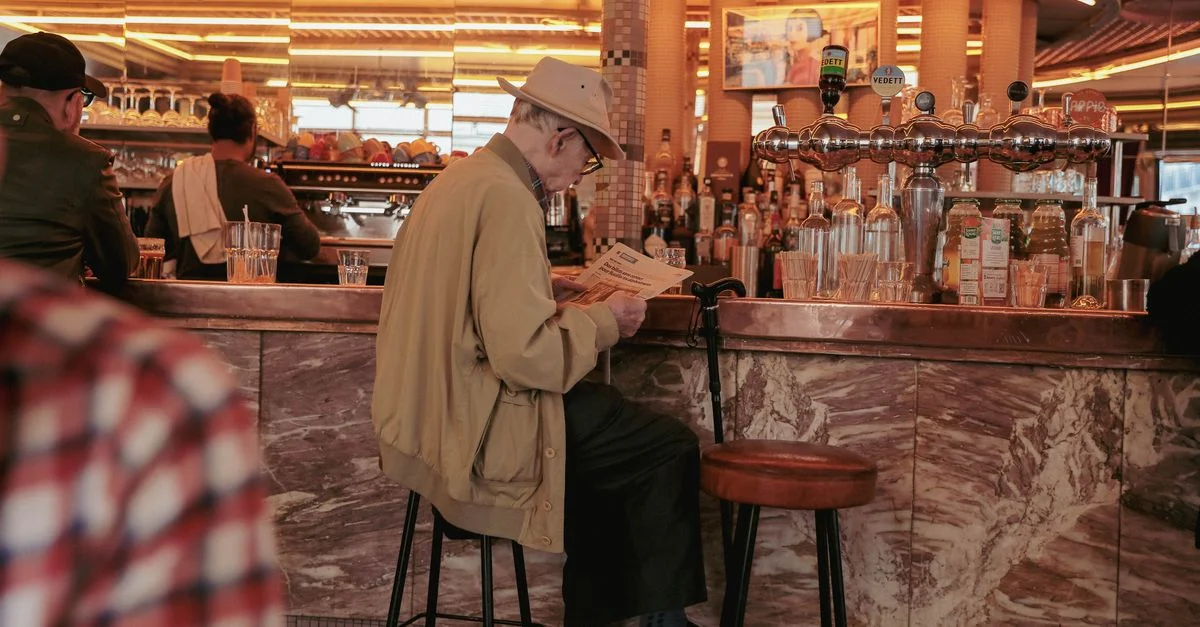 Un homme senior avec un chapeau lit un journal dans un bar, capturant un moment paisible à l'intérieur.