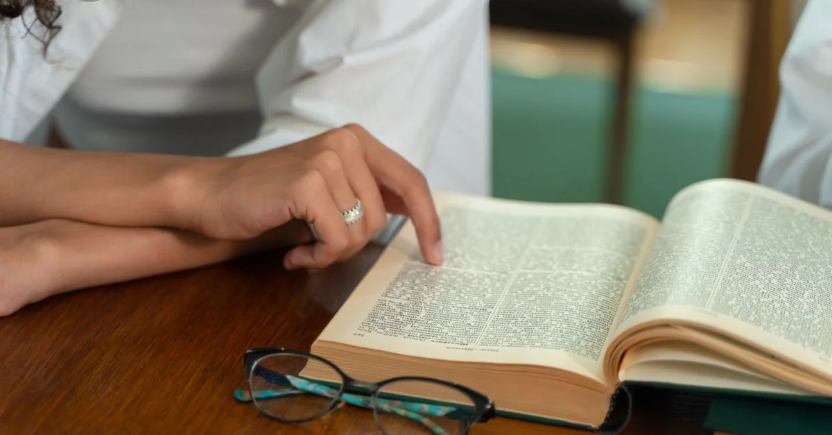 Gros plan sur une personne lisant un livre ouvert avec des lunettes sur un bureau en bois.